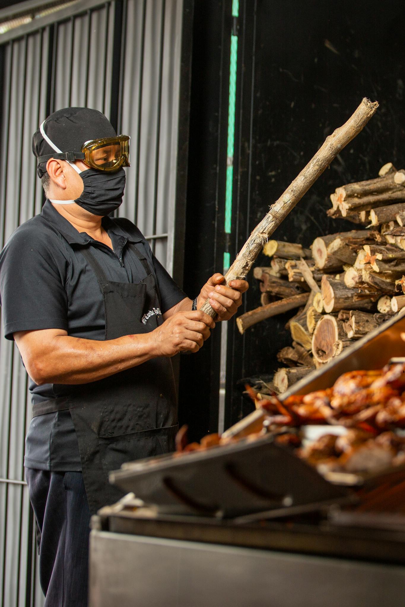 Masked chef preparing a barbecue using firewood at an outdoor grill, enhancing flavor with traditional methods.