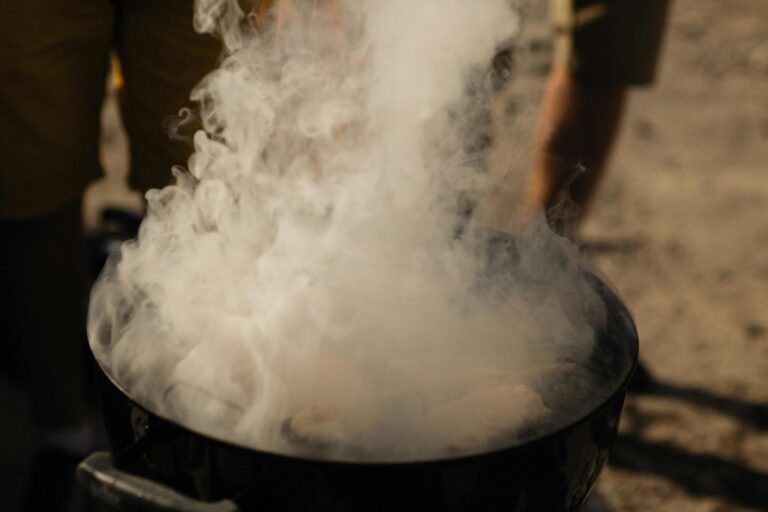 A smoky grill at an outdoor barbecue with people preparing food in a casual setting.